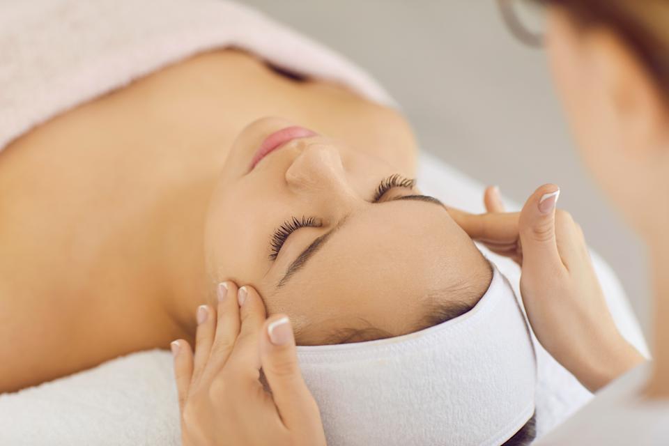 Relaxed woman relieves stress while receiving professional facial and head massage in spa salon. Close up of female masseur hands doing massage to client lying with eyes closed in white bandage.